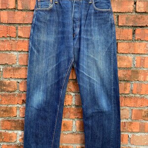 Puede incluir: Vaqueros de mezclilla azul vintage con bragueta de botones y diseño clásico de cinco bolsillos. Los vaqueros muestran signos de desgaste y decoloración, dándoles un aspecto desgastado. Los vaqueros cuelgan de una percha dorada contra una pared de ladrillos.
