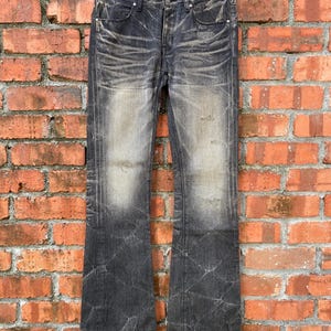 May include: Faded black denim jeans with a distressed, worn appearance. The jeans have a bootcut style, with a lighter wash on the front and back. They are hanging on a wooden hanger against a brick wall.