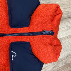 May include: A black and orange puffer jacket with a zipper closure. The jacket has a high collar and a patch pocket on the left side.