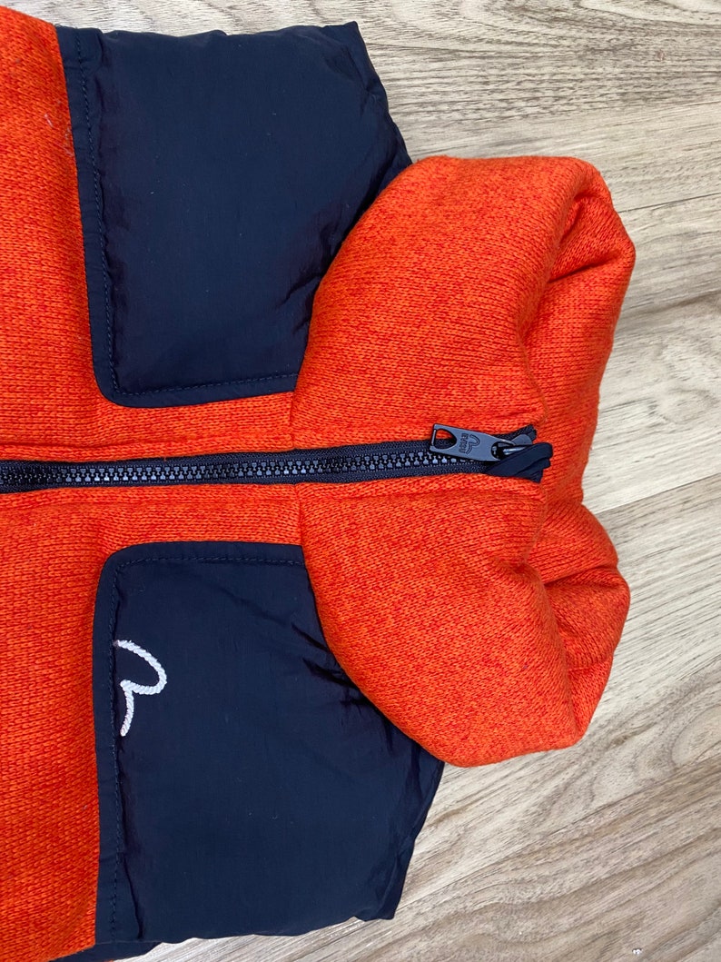 May include: A black and orange puffer jacket with a zipper closure. The jacket has a high collar and a patch pocket on the left side.