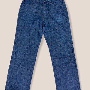 May include: Blue denim jeans with a unique patchwork design. The jeans have a classic five-pocket style, a button and zipper closure, and a straight leg fit. The patchwork creates a grid-like pattern across the front and back.