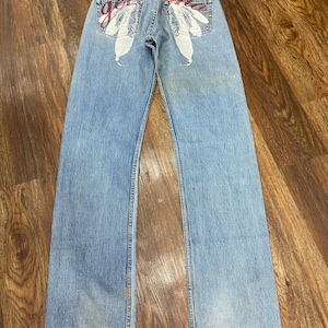 Puede incluir: Un par de pantalones vaqueros azules con un diseño gráfico blanco y rojo en los bolsillos traseros. El diseño gráfico presenta plumas y la palabra "Genuine" en rojo.