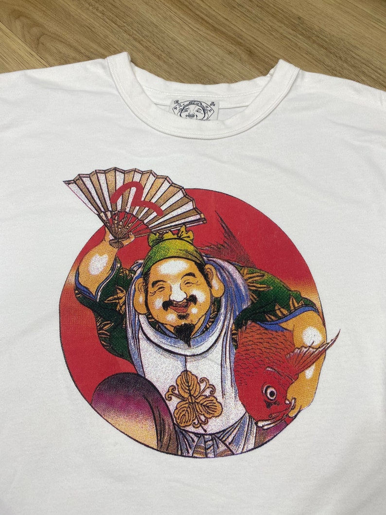 May include: White t-shirt with a red circle graphic featuring a cartoon character holding a fan and a fish. The character is wearing a green hat and a white and blue outfit. The text "福" is visible on the graphic.