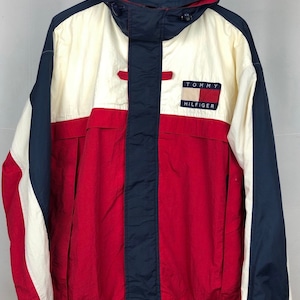 May include: A red, white, and navy blue windbreaker jacket with a Tommy Hilfiger logo on the chest. The jacket has a hood and a zipper closure.
