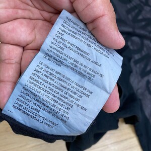 May include: A white care tag with black text detailing the care instructions for a garment. The text reads: "This fabric is foil transfer. Decoloration and peeling will occur with friction due to wear and cleaning. The fabric is sensitive to heat. Handle with care. The print is not permanent. Color will run with friction. Handle with care. Hand wash in a net bag. Do not bleach. Do not tumble dry. Line dry in the shade. Wash colored garments separately. Do not steam."