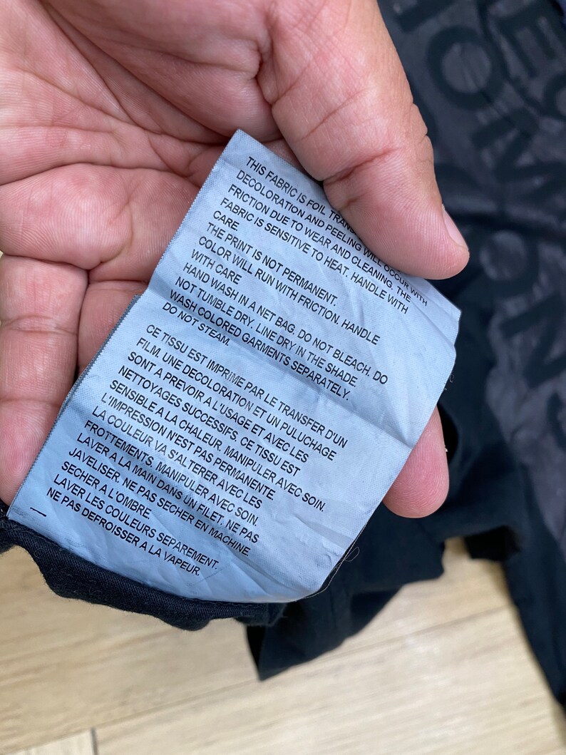 May include: A white care tag with black text detailing the care instructions for a garment. The text reads: "This fabric is foil transfer. Decoloration and peeling will occur with friction due to wear and cleaning. The fabric is sensitive to heat. Handle with care. The print is not permanent. Color will run with friction. Handle with care. Hand wash in a net bag. Do not bleach. Do not tumble dry. Line dry in the shade. Wash colored garments separately. Do not steam."