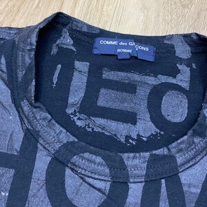 May include: Black t-shirt with a large, faded print of the words "Comme des Gar&ccedil;ons Homme" on the front. The print is in a distressed, layered style.