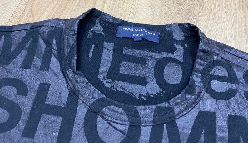 May include: Black t-shirt with a large, faded print of the words "Comme des Gar&ccedil;ons Homme" on the front. The print is in a distressed, layered style.