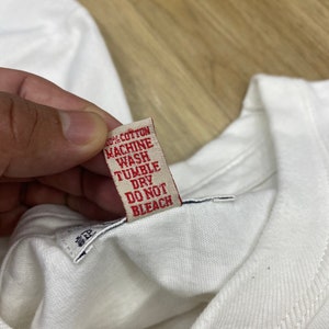 May include: A white cotton t-shirt with a red care label that reads "100% Cotton, Machine Wash, Tumble Dry, Do Not Bleach".