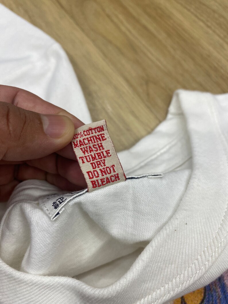 May include: A white cotton t-shirt with a red care label that reads "100% Cotton, Machine Wash, Tumble Dry, Do Not Bleach".