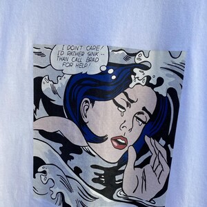 May include: White t-shirt featuring a comic book style graphic of a woman with blue hair crying in water. The speech bubble reads, "I DON'T CARE! I'D RATHER SINK -- THAN CALL BRAD FOR HELP!"