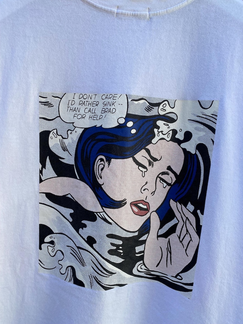 May include: White t-shirt featuring a comic book style graphic of a woman with blue hair crying in water. The speech bubble reads, "I DON'T CARE! I'D RATHER SINK -- THAN CALL BRAD FOR HELP!"