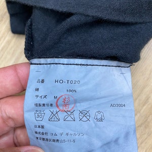 May include: A blue clothing tag with black text and symbols. The tag says "HO-T020", "100%", "Size M", "Made in Japan", and "Commes des Garcons" with an address in Tokyo. The tag also includes care instructions.