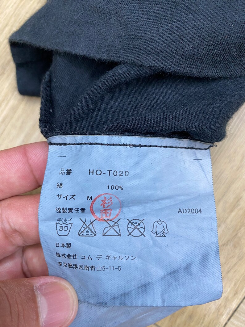 May include: A blue clothing tag with black text and symbols. The tag says "HO-T020", "100%", "Size M", "Made in Japan", and "Commes des Garcons" with an address in Tokyo. The tag also includes care instructions.