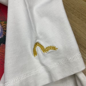 May include: White t-shirt with a gold embroidered design of wheat stalks on the sleeve.