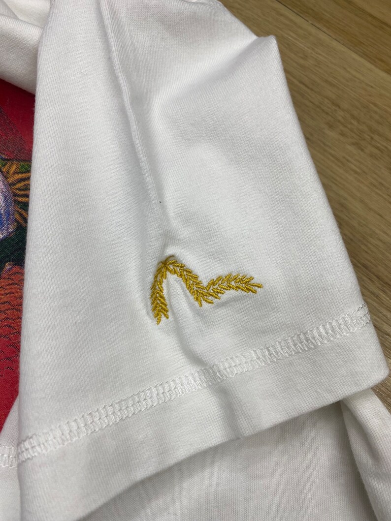May include: White t-shirt with a gold embroidered design of wheat stalks on the sleeve.