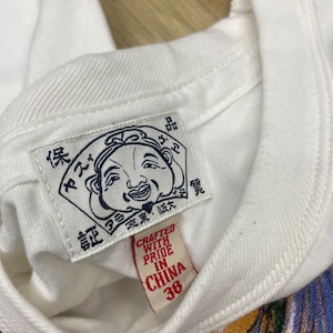May include: White t-shirt with a red tag that says "Crafted with Pride in China 38". The tag is attached to a white label with a cartoon illustration of a smiling man with a large nose and a red hat. The label has Japanese text.