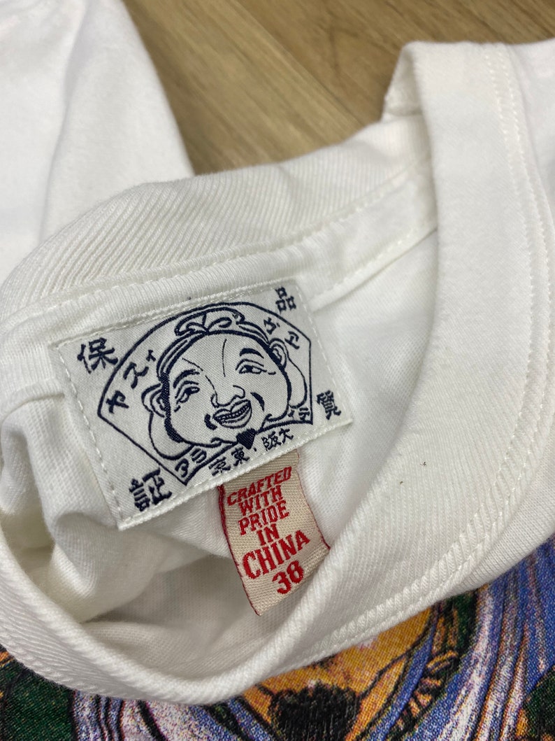 May include: White t-shirt with a red tag that says "Crafted with Pride in China 38". The tag is attached to a white label with a cartoon illustration of a smiling man with a large nose and a red hat. The label has Japanese text.