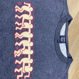 May include: A dark gray crew neck sweatshirt featuring a stacked figure design in cream and red. The design is a repeating pattern of stylized human figures. The sweatshirt has a ribbed neckline and a tag on the inside.