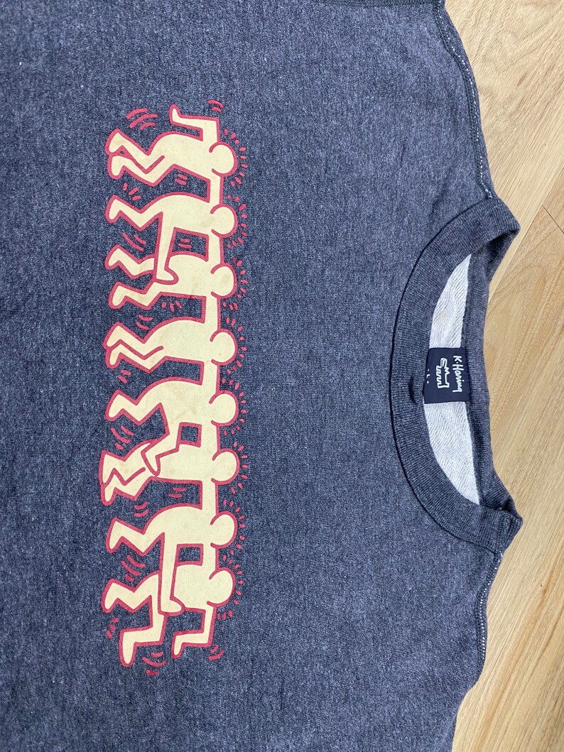 May include: A dark gray crew neck sweatshirt featuring a stacked figure design in cream and red. The design is a repeating pattern of stylized human figures. The sweatshirt has a ribbed neckline and a tag on the inside.