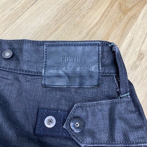 Peut inclure: Jean en denim noir avec un patch en cuir sur lequel est inscrit "EDWIN E-FUNCTION" et "STRETCH DENIM" en blanc.