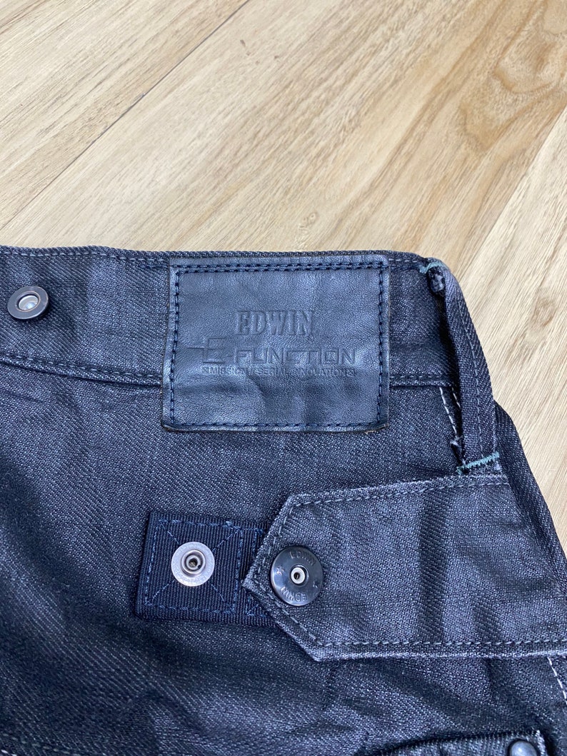 Peut inclure: Jean en denim noir avec un patch en cuir sur lequel est inscrit "EDWIN E-FUNCTION" et "STRETCH DENIM" en blanc.