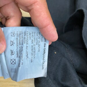 May include: A white tag with black text attached to a black garment. The tag reads "COMME des GAR&Ccedil;ONS Co., Ltd. -T020 MADE IN JAPAN 100% COTTON HAND WASH. DO NOT BLEACH. DO NOT IRON. DO NOT DRY CLEAN. LINE DRY. FABRIQUE AU JAPON 100% COTON LAVAGE MAINS. JAVEL INTERDIT. NE PAS REPASSER. INTERDIT NETTOYAGE A SEC. SECHAGE A L'OMBRE."