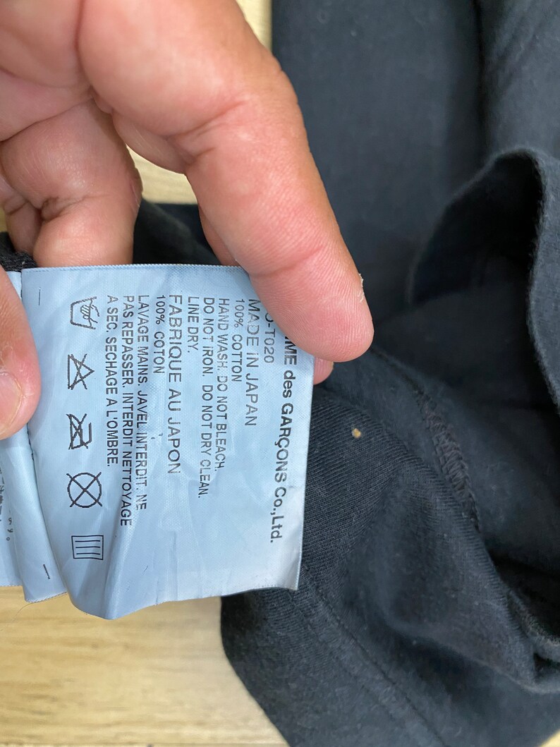 May include: A white tag with black text attached to a black garment. The tag reads "COMME des GAR&Ccedil;ONS Co., Ltd. -T020 MADE IN JAPAN 100% COTTON HAND WASH. DO NOT BLEACH. DO NOT IRON. DO NOT DRY CLEAN. LINE DRY. FABRIQUE AU JAPON 100% COTON LAVAGE MAINS. JAVEL INTERDIT. NE PAS REPASSER. INTERDIT NETTOYAGE A SEC. SECHAGE A L'OMBRE."