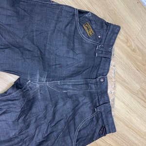 Peut inclure: Jean en denim gris fonc&eacute; avec un petit patch jaune sur la poche droite qui dit "G-Star RAW Denim Supply Co. 1989 Amsterdam".
