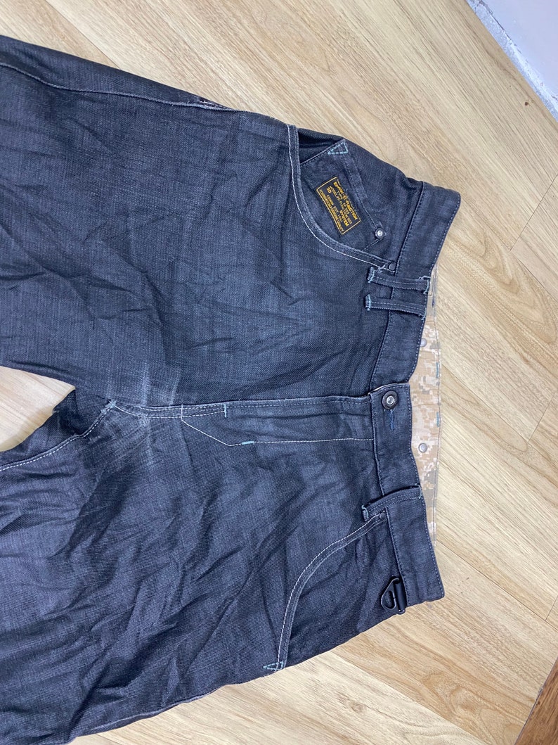 Peut inclure: Jean en denim gris fonc&eacute; avec un petit patch jaune sur la poche droite qui dit "G-Star RAW Denim Supply Co. 1989 Amsterdam".