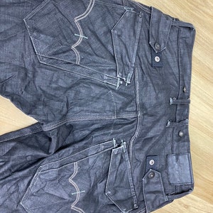 Peut inclure: Un jean en denim gris fonc&eacute; avec plusieurs poches et des d&eacute;tails de couture. Le jean a un aspect d&eacute;lav&eacute; et une &eacute;tiquette de marque sur la poche arri&egrave;re droite.