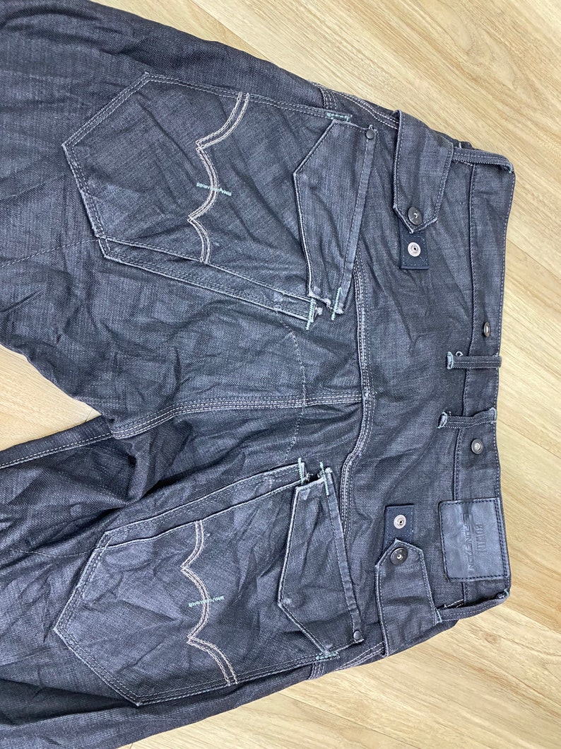 Peut inclure: Un jean en denim gris fonc&eacute; avec plusieurs poches et des d&eacute;tails de couture. Le jean a un aspect d&eacute;lav&eacute; et une &eacute;tiquette de marque sur la poche arri&egrave;re droite.
