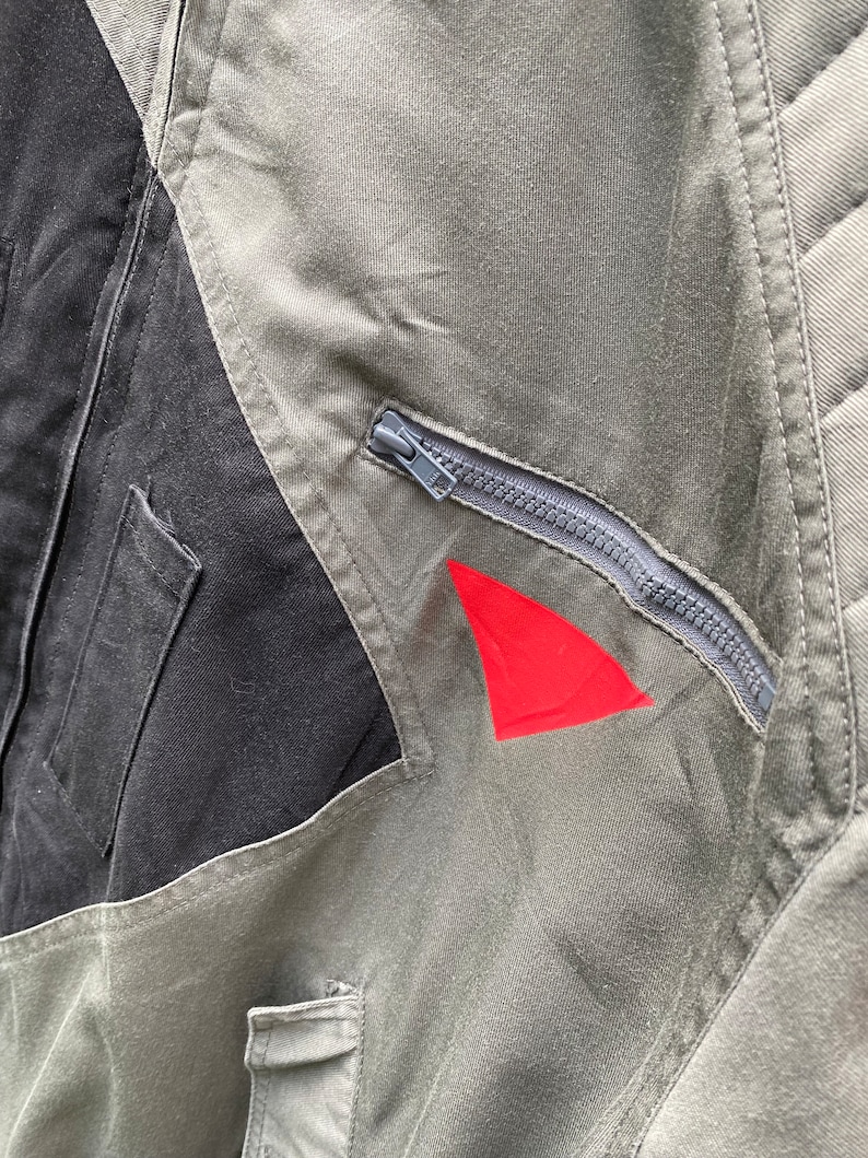 May include: A close-up of a green and black jacket with a zipper and a red triangle patch. The jacket has a quilted design on the back.