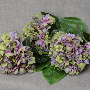 May include: A bouquet of three artificial hydrangea flowers with green leaves. The flowers are a mix of light green and purple.