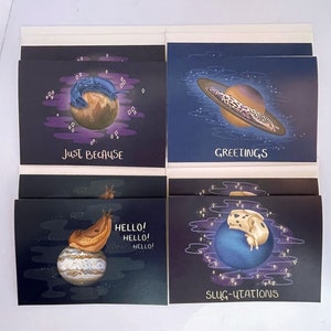 May include: A set of four greeting cards featuring quirky illustrations of slugs on planets. Each card has a unique design with text: "Just Because", "Greetings", "Hello!", and "Slug-utations". The cards have a dark blue background with purple accents.