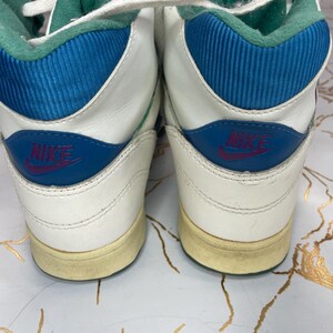 Puede incluir: Un par de zapatillas blancas de ca&ntilde;a alta con detalles azules y verdes. Las zapatillas tienen un logotipo de Nike en el lateral y una suela blanca.