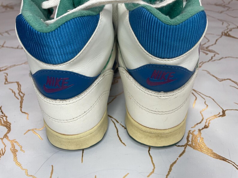 Puede incluir: Un par de zapatillas blancas de ca&ntilde;a alta con detalles azules y verdes. Las zapatillas tienen un logotipo de Nike en el lateral y una suela blanca.