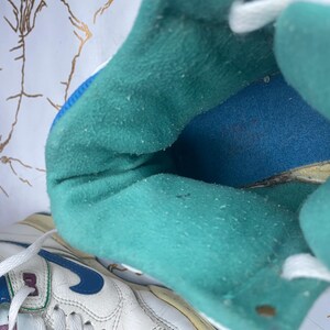 Puede incluir: Primer plano del interior de una zapatilla blanca con forro azul turquesa y leng&uuml;eta blanca. La zapatilla tiene suela blanca y un logotipo Swoosh azul.