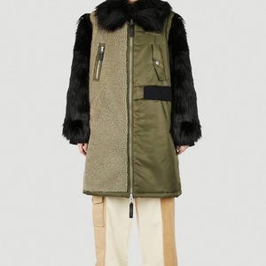 Peut inclure: Un long manteau vert olive et beige avec des manches et un col en fausse fourrure noire. Le manteau a une fermeture éclair et une texture contrastée sur le devant. Le modèle porte un pantalon beige et crème et des chaussures noires.