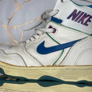 Puede incluir: Zapatillas blancas de ca&ntilde;a alta con un swoosh Nike azul y detalles verdes. La zapatilla tiene una parte superior de cuero blanco y una suela de goma blanca. El logotipo de Nike est&aacute; impreso en el lateral de la zapatilla en rosa.