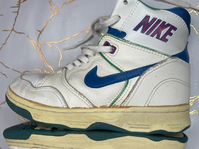 Puede incluir: Zapatillas blancas de ca&ntilde;a alta con un swoosh Nike azul y detalles verdes. La zapatilla tiene una parte superior de cuero blanco y una suela de goma blanca. El logotipo de Nike est&aacute; impreso en el lateral de la zapatilla en rosa.