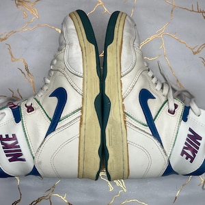 Puede incluir: Un par de zapatillas blancas de ca&ntilde;a alta con detalles en azul y verde. Las zapatillas tienen la palabra "NIKE" escrita en el lateral en morado.