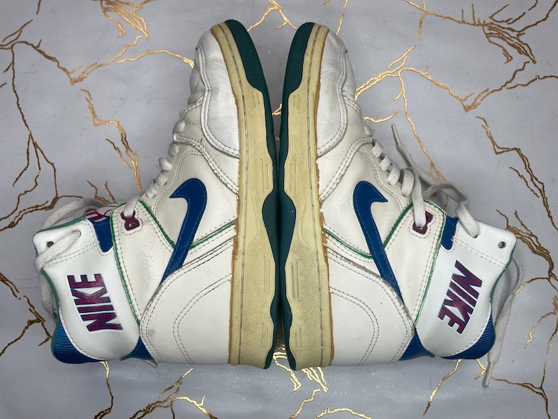 Puede incluir: Un par de zapatillas blancas de ca&ntilde;a alta con detalles en azul y verde. Las zapatillas tienen la palabra "NIKE" escrita en el lateral en morado.