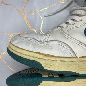 Puede incluir: Zapatillas blancas de cuero de ca&ntilde;a alta con un logotipo Swoosh azul y una suela azul verdosa y blanca.