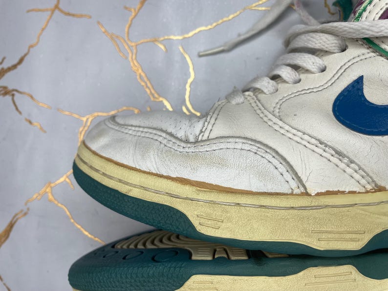 Puede incluir: Zapatillas blancas de cuero de ca&ntilde;a alta con un logotipo Swoosh azul y una suela azul verdosa y blanca.