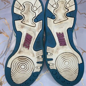 Puede incluir: Zapatillas blancas de cuero con suela exterior azul turquesa y blanca. Las zapatillas tienen un logotipo de Nike rosa en la parte posterior.