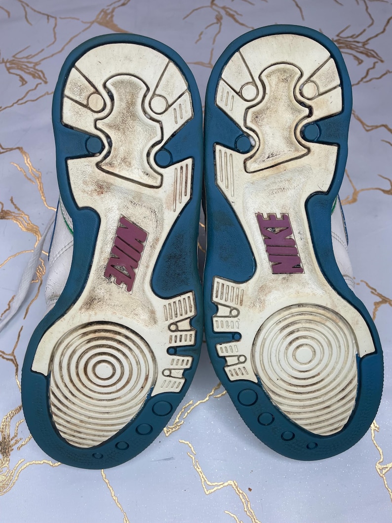 Puede incluir: Zapatillas blancas de cuero con suela exterior azul turquesa y blanca. Las zapatillas tienen un logotipo de Nike rosa en la parte posterior.