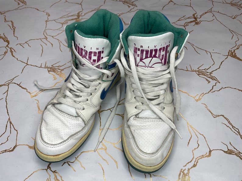 Puede incluir: Un par de zapatillas blancas de ca&ntilde;a alta con detalles en azul y verde. Las zapatillas tienen el texto "Driving Force" en el lateral.