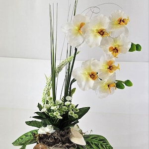 May include: A white orchid arrangement in a gray pot with a heart design. The arrangement includes green leaves and a small brown tag that says "Hand made".