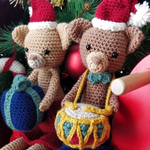 May include: Two crocheted teddy bears wearing Santa hats, one holding a blue and white gift box and the other holding a yellow and green drum. The bears are sitting on a red chair.